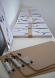 A series of clipboards are arranged in a row on a white surface. Sheets of paper with abstract black and white designs are neatly stacked and secured with brown paper bands. The scene gives a minimalist and organized look.