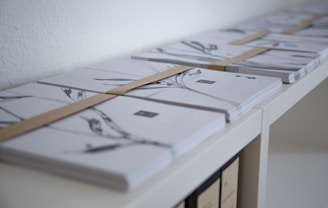 Stacks of neatly bound papers or envelopes are arranged on a white shelf. Each stack is secured with a brown band, and the items appear to have minimalistic black and white designs on their surfaces.