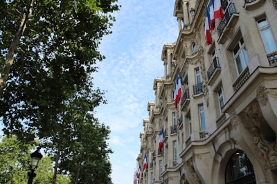 An elegant building with ornate architectural details is adorned with multiple French flags. The building is situated next to a street lined with lush green trees. The sky is mostly clear with scattered white clouds.