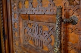 Intricate and ornate wooden door featuring detailed geometric patterns and carvings, with a decorative metal handle. The wood is richly stained, showcasing its natural grain.