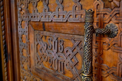 Intricate and ornate wooden door featuring detailed geometric patterns and carvings, with a decorative metal handle. The wood is richly stained, showcasing its natural grain.
