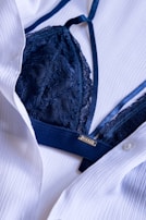 A dark blue lace bra is partially covered by a white button-up shirt. The fabric of both the bra and the shirt appears to be of fine texture, with the bra having intricate lace details and the shirt having a smooth, slightly crinkled texture.