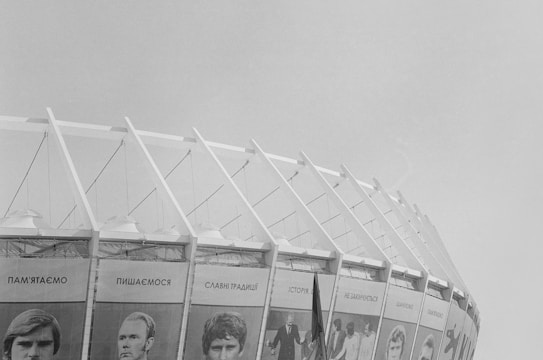 A large, modern stadium structure with distinctive slanted beams and large banners featuring black and white portraits. The banners include text in Cyrillic script. The overall atmosphere is open and minimalistic against a clear sky.