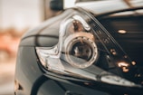 A close-up view of a sleek modern car headlight with a reflective and metallic finish. The image captures the intricate details of the headlamp, including the circular design and the smooth contours of the car body. The background is softly blurred, emphasizing the focus on the headlight.