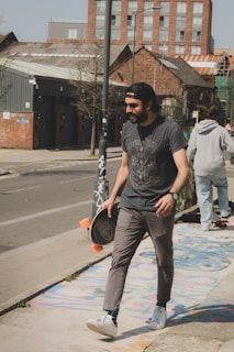 A man is walking down a urban street holding a skateboard by his side. He is wearing a black cap, a dark t-shirt, gray pants, and white sneakers. The background features industrial-style buildings with brick facades and some trees, suggesting an urban environment. Another person in casual clothing is seen in the background.