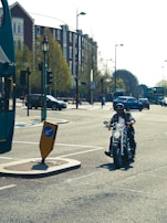 A motorcyclist wearing a helmet rides along a city street. Nearby, a double-decker bus is partially visible, and several cars can be seen on the road. The scene is set in an urban environment with brick buildings and trees along the street.