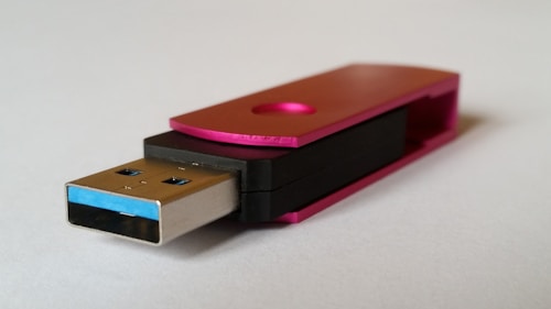 A USB flash drive with a metal connector and a pink metallic cap. The body is partially black with a simple, modern design. The flash drive is resting on a neutral, light-colored surface.