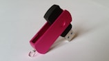 A pink USB flash drive with a swivel cover is positioned with the metal connector exposed. The cover has a small circular indent for grip and a metal loop for attachment. The background is plain and light-colored.