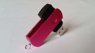 A pink USB flash drive with a swivel cover is positioned with the metal connector exposed. The cover has a small circular indent for grip and a metal loop for attachment. The background is plain and light-colored.