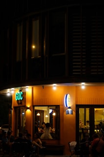A small café or restaurant with an inviting orange exterior, illuminated by several lights. People are seated inside and outside, enjoying a relaxed evening atmosphere. The signage reads 'GOC' in bright green letters.
