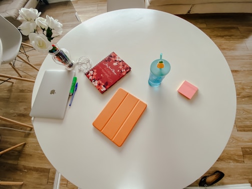 A small round white table with various items arranged neatly on top: a closed silver laptop with a pen and highlighter beside it, a red floral notebook with earbuds, an orange tablet or notebook, a blue cup with a straw and a pineapple decoration, and a stack of pink sticky notes. There is also a vase of white flowers on a chair and a pair of black shoes on the floor nearby.