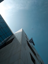 A low-angle view of a modern building with a geometric, triangular shape against a bright sky. The building features a prominent Greek flag flying from one corner, and a single bird is visible in the sky.