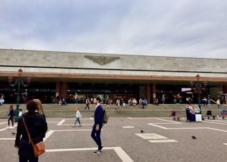 A large, rectangular building with a modern facade, featuring a logo in the center. People are gathered in front of the building, some walking while others are sitting on steps. The area is paved and spacious, with street lamps and a few pigeons visible.