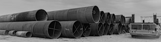 A series of large industrial metal pipes stacked horizontally in three rows on a gravel surface. Beside the pipes, on the right, is a heavy-duty industrial vehicle with a flatbed. The sky appears overcast, casting a monochromatic tone over the scene.
