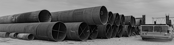 A series of large industrial metal pipes stacked horizontally in three rows on a gravel surface. Beside the pipes, on the right, is a heavy-duty industrial vehicle with a flatbed. The sky appears overcast, casting a monochromatic tone over the scene.