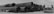 A series of large industrial metal pipes stacked horizontally in three rows on a gravel surface. Beside the pipes, on the right, is a heavy-duty industrial vehicle with a flatbed. The sky appears overcast, casting a monochromatic tone over the scene.