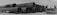 A series of large industrial metal pipes stacked horizontally in three rows on a gravel surface. Beside the pipes, on the right, is a heavy-duty industrial vehicle with a flatbed. The sky appears overcast, casting a monochromatic tone over the scene.