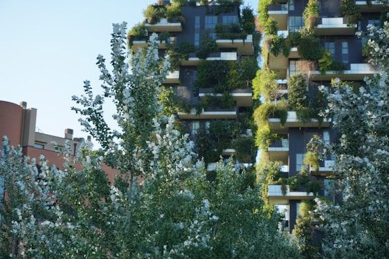 Vertical gardens adorn the facade of a modern building, blending architecture with lush greenery. Several balconies are heavily planted with a variety of shrubs and trees, creating an urban oasis. In the foreground, leafy trees partially obscure the view, adding to the overall green aesthetic.