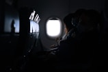 The interior of an airplane cabin with passengers seated. The focus is on a woman looking out the window while wearing a mask. Another individual is also visible, resting or sleeping, with a mask on. The lighting is dim, highlighting the window and creating a quiet and subdued atmosphere.