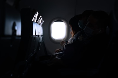 The interior of an airplane cabin with passengers seated. The focus is on a woman looking out the window while wearing a mask. Another individual is also visible, resting or sleeping, with a mask on. The lighting is dim, highlighting the window and creating a quiet and subdued atmosphere.