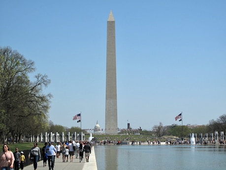 A tall stone monument with a pointed top stands prominently in the center, surrounded by a gathering of people. Trees flank the sides of a water body reflecting the clear blue sky above. American flags wave gently, adding a patriotic element to the scene.