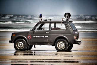 A rugged off-road vehicle parked on a sandy beach nearby the ocean, equipped with various communication and navigational devices on its roof. The vehicle has signage, including the words 'Ville de Soulac/Mer,' suggesting it might be used for official purposes related to the beach or town. The background shows waves crashing against the shore, indicating a windy or active sea.