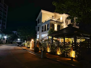 A well-lit house during nighttime with large windows partially covered by curtains. The structure is multi-story, surrounded by a wooden fence partially covered with plants. The foreground features a patio area with additional lighting and greenery, and the background has trees lining the street.