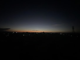 A dark horizon where the last light of the day is disappearing. The sky gradates from a deep blue at the top to a faint orange near the horizon. Distant city lights are faintly visible, and a tall communication tower stands to the right.
