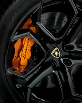 A close-up view of an elegant black alloy wheel featuring a prominent orange brake caliper with Lamborghini branding. The intricate design and sheen of the wheel highlight the luxury and craftsmanship.