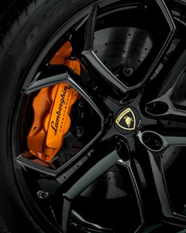 A close-up view of an elegant black alloy wheel featuring a prominent orange brake caliper with Lamborghini branding. The intricate design and sheen of the wheel highlight the luxury and craftsmanship.