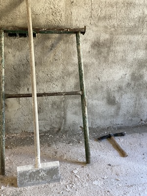 A rusty metal ladder leans against a textured concrete wall. On the ground, there is a hammer lying next to some scattered dust and debris, as well as a tool with a flat, wide blade on a wooden handle.