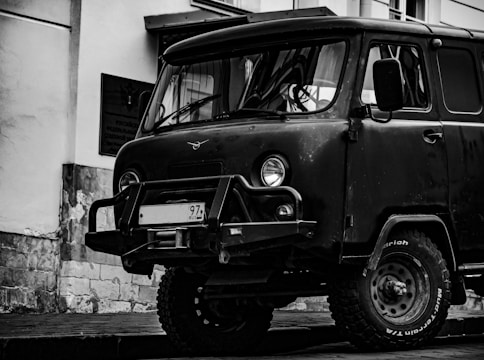 A vintage, rugged van with off-road tires is parked beside a building with stone and plaster walls. The front grill and bumper are prominent, and the van's design appears to be utilitarian and robust.