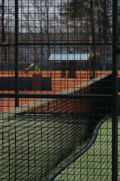 A tennis court is surrounded by a green wire mesh fence. The net in the center of the court divides the space, while the background shows blurred trees and possibly another court area. The ground is covered with artificial grass, and the scene is quiet and still.