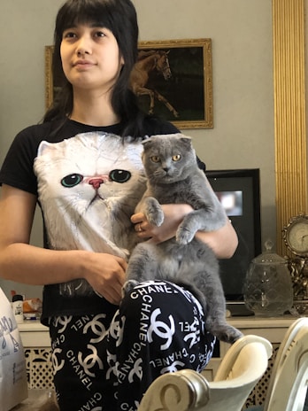 A person is standing indoors, holding a gray cat. The person is wearing a t-shirt with a large cat face printed on it. Behind them, a wall decorated with an ornate gold frame contains a painting of a horse. A vintage clock and decorative glassware are visible in the background.