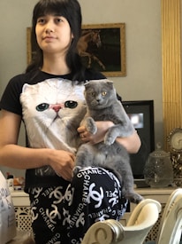 A person is standing indoors, holding a gray cat. The person is wearing a t-shirt with a large cat face printed on it. Behind them, a wall decorated with an ornate gold frame contains a painting of a horse. A vintage clock and decorative glassware are visible in the background.
