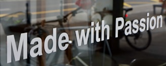 The image features a glass window with the phrase 'Made with Passion' prominently displayed in white text. Behind the glass, there is a blurred view of a street scene, including a bicycle and some indistinct urban elements.