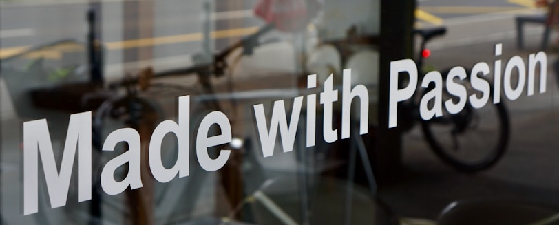 The image features a glass window with the phrase 'Made with Passion' prominently displayed in white text. Behind the glass, there is a blurred view of a street scene, including a bicycle and some indistinct urban elements.
