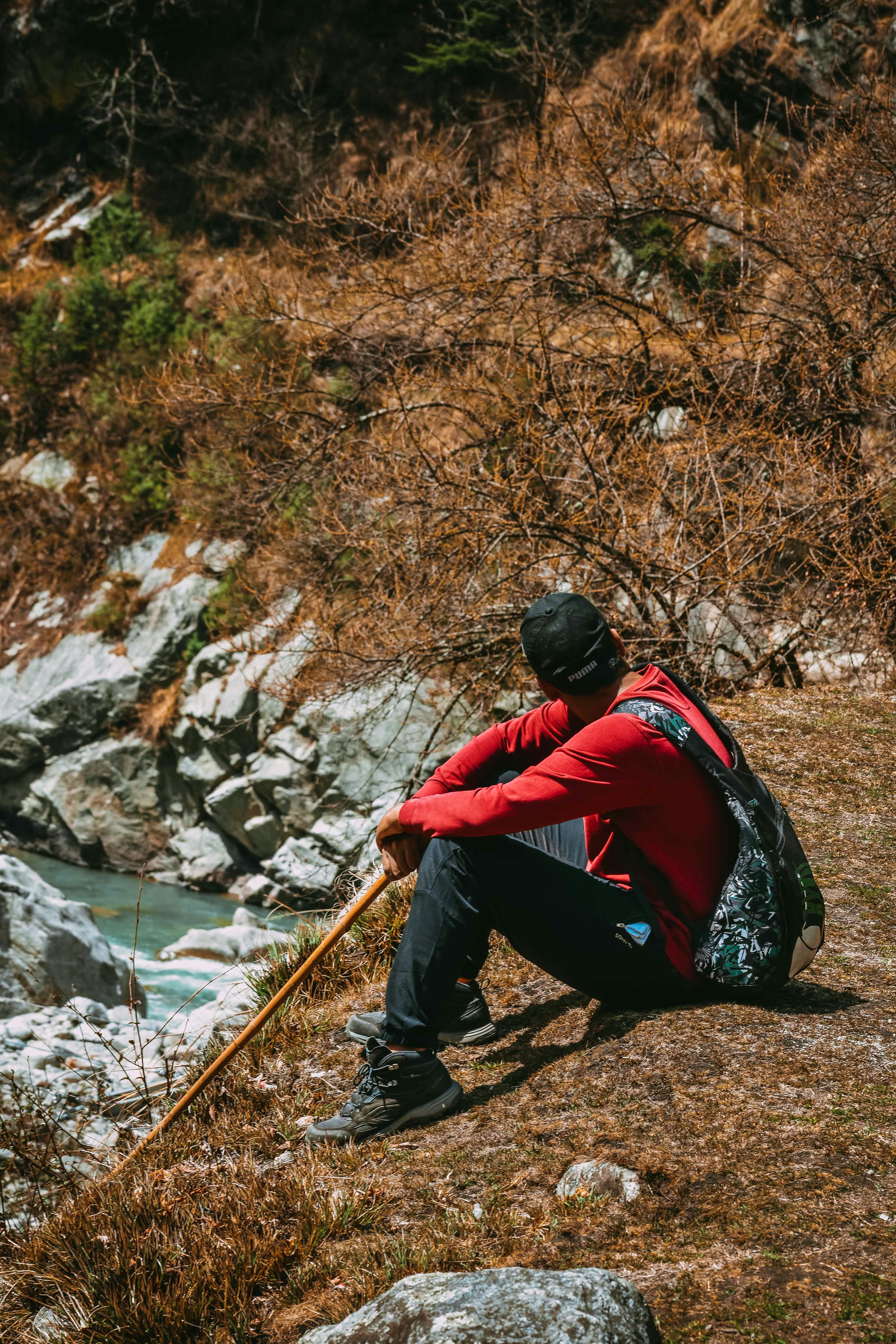 man in red and black jacket sitting on rock near river during daytime
