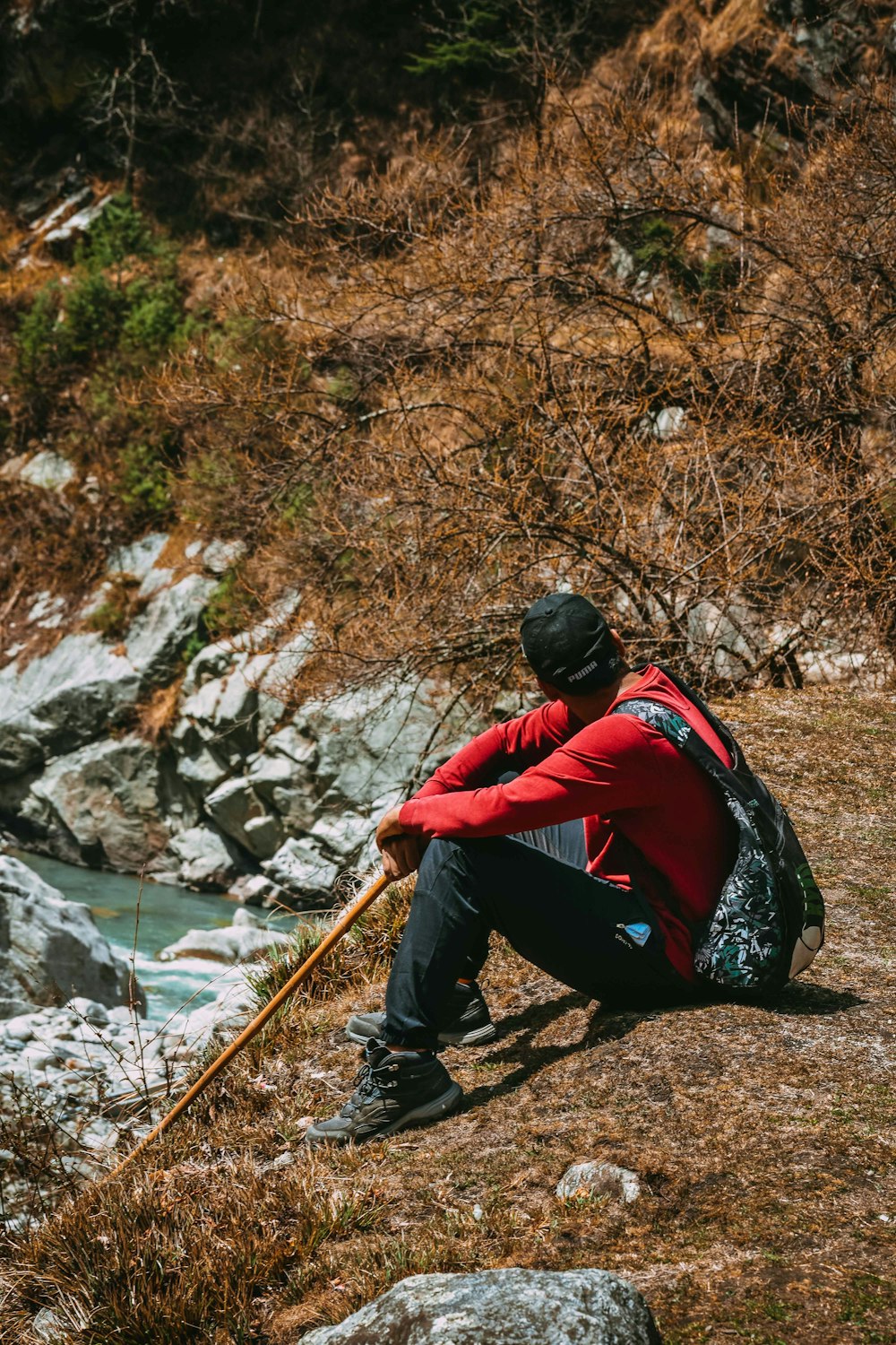 낮에 강 근처 바위에 앉아 있는 빨간색과 검은색 재킷을 입은 남자