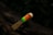 A close-up view of a wooden toothbrush with brightly colored bristles including green, yellow, blue, and red. The background is natural and blurred, possibly depicting a tree branch or dark foliage.