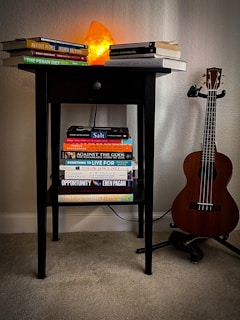 A small black table is set against a light-colored wall with a soft glow emitting from an orange Himalayan salt lamp placed on top. Several books are stacked on the tabletop and a lower shelf, with titles visible across different genres. To the right of the table, a wooden ukulele is positioned on a stand.