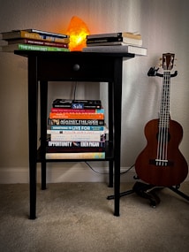A small black table is set against a light-colored wall with a soft glow emitting from an orange Himalayan salt lamp placed on top. Several books are stacked on the tabletop and a lower shelf, with titles visible across different genres. To the right of the table, a wooden ukulele is positioned on a stand.