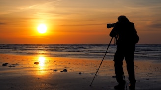 A photographer is silhouetted against a vibrant sunset over the ocean, standing on the beach with a camera and tripod. The sun is low in the sky, creating a golden reflection on the wet sand and waves.