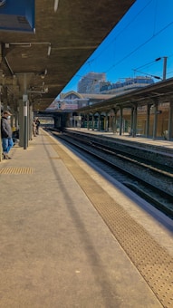 A railway platform with multiple tracks, overhead structures, and a few people waiting. In the background, a city skyline with residential buildings is visible under a clear blue sky. The platform is clean and well-maintained, with tactile paving along the edge.