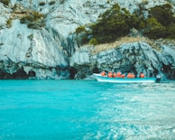 A boat with several people wearing orange life jackets is situated on a clear turquoise body of water. In the background, there are rugged rock formations with patches of greenery growing on them.
