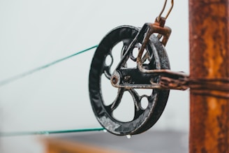 A close-up view of a metal pulley attached to a rusted post. The pulley appears weathered, with visible signs of rust and wear. Two green cables run through the pulley, contrasting with the dark color of the metal.