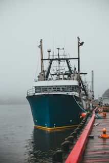 A large fishing boat is docked at a pier on a foggy day. The boat is painted with dark blue and white colors, equipped with various fishing equipment and antennas. The water is calm, and there is a red railing along the pier. Some small fishing supplies and ropes are scattered on the wooden pier.