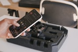 A person is holding a smartphone displaying a social media app, while a black foam case containing a compact gimbal stabilizer is on the table. In the background, there is a chair with a white frame and a wooden desk with various items on it.