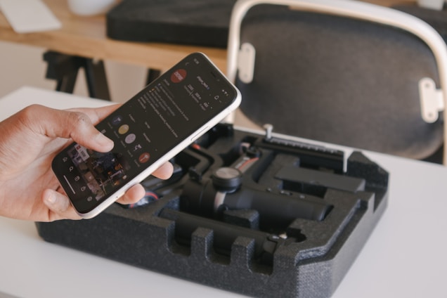 A person is holding a smartphone displaying a social media app, while a black foam case containing a compact gimbal stabilizer is on the table. In the background, there is a chair with a white frame and a wooden desk with various items on it.