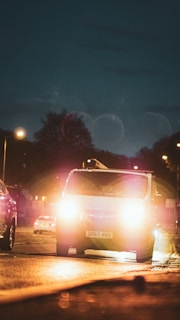 A van with headlights on is prominent in the foreground, driving on a dimly lit street during the night. Surrounding the van, other vehicles are visible, and streetlights illuminate parts of the road. The background shows a dark sky and silhouettes of trees.